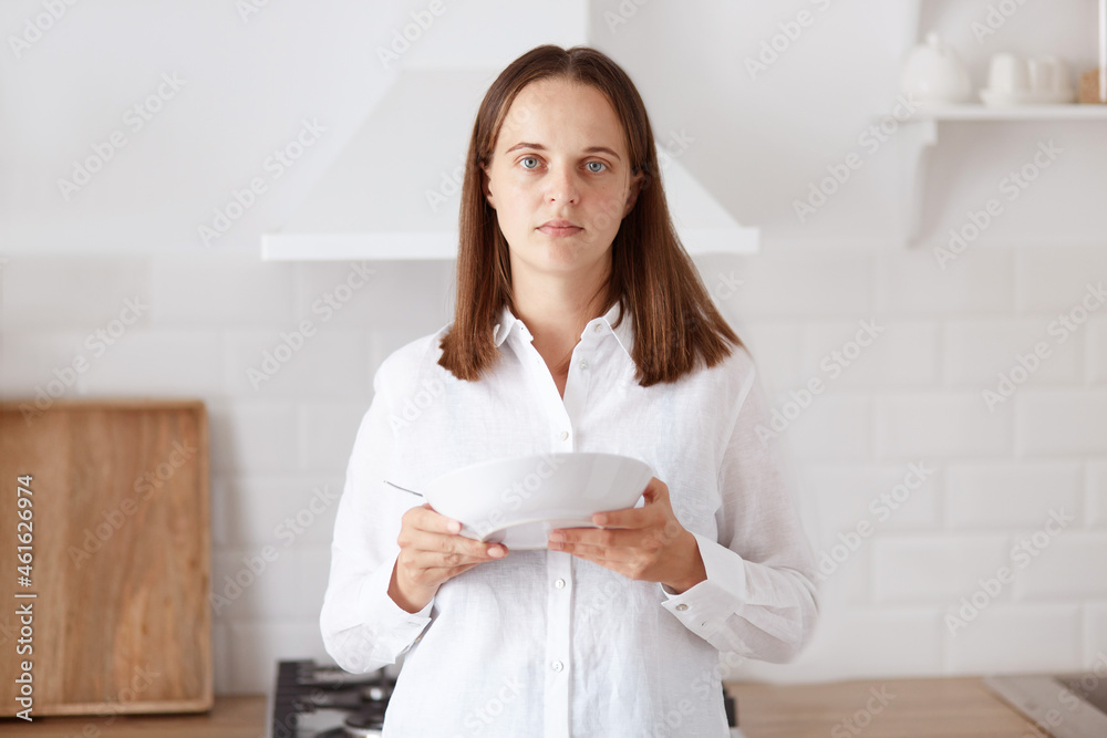 Sad upset attractive young adult woman with dark hair wearing white casual shirt, having breakfast, holding plate in hands, looking at away, eating from spoon. Sad upset attractive young adult woman with dark hair wearing white casual shirt, having breakfast, holding plate in hands, looking at away, eating from spoon.