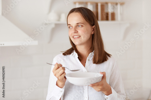 Portrait of happy smiling young adult woman eating from plate in the morning, looking away with positive expression, posing with kitchen set on background.