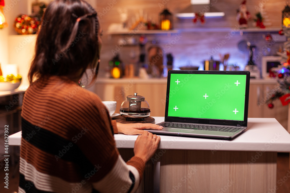Modern young adult watching green screen on laptop digital device sitting at festive counter kitchen. Woman using computer chroma key gadget on background display for mockup template
