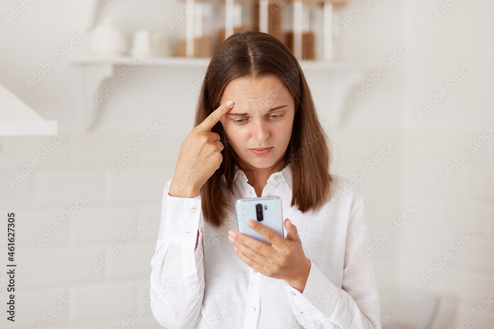 Confused puzzled dark haired woman standing cell phone in hands, checking social networks, reading negative message or comment, posing in the kitchen at home. Confused puzzled dark haired woman standing cell phone in hands, checking social networks, reading negative message or comment, posing in the kitchen at home.
