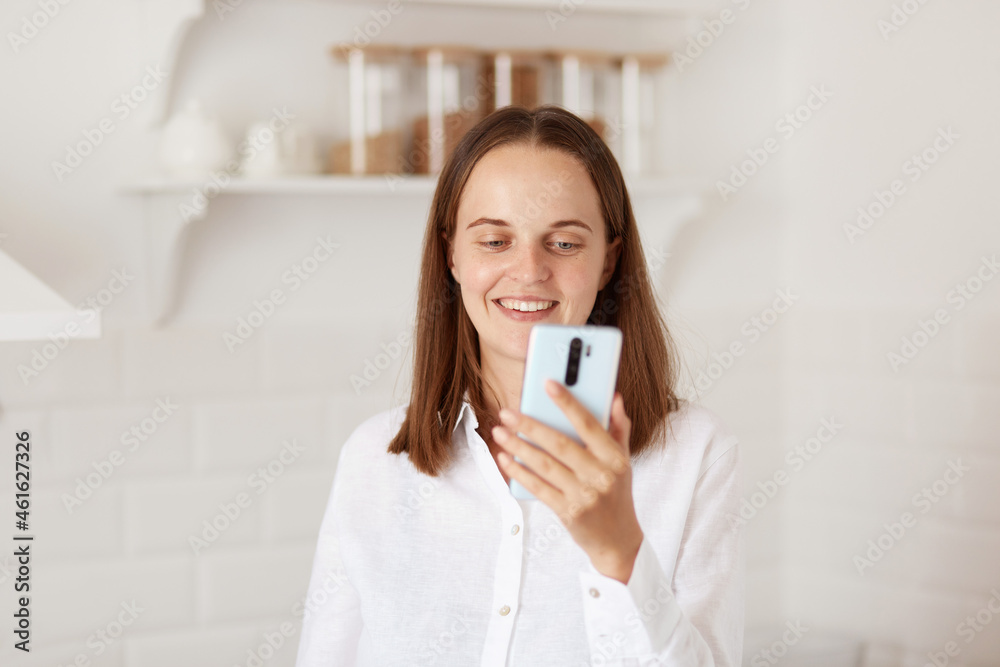 Indoor shot of positive smiling female with dark hair, looking at cell phone in hands, broadcasting livestream or having video call, wearing casual clothing, posing in kitchen. Indoor shot of positive smiling female with dark hair, looking at cell phone in hands, broadcasting livestream or having video call, wearing casual clothing, posing in kitchen.