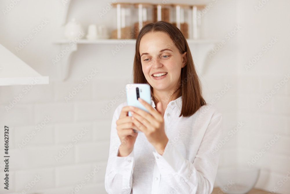 Optimistic smiling young adult woman with dark hair holding cell phone in hands, having video call or girl blogger talking with her followers online, posing in kitchen. Optimistic smiling young adult woman with dark hair holding cell phone in hands, having video call or girl blogger talking with her followers online, posing in kitchen.