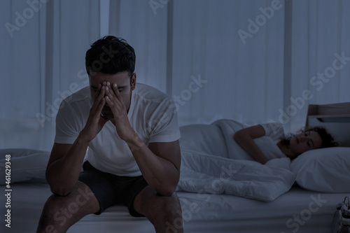 Upset depressed young man sitting on the edge of bed against his wife lying on the bed. Relationship difficulties , Problems couple.