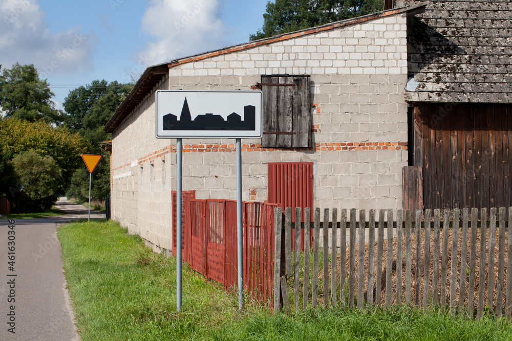 A road sign indicating a built-up area with a background of rural farm buildings. A road sign indicating a built-up area with a background of rural farm buildings.