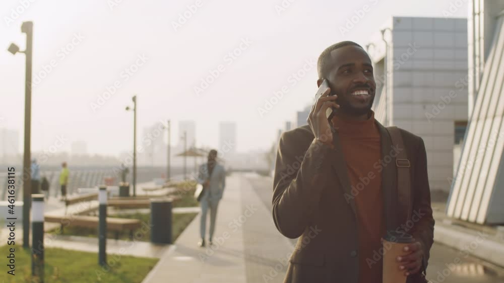 Young positive African American businessman walking on street in city, holding to go coffee and speaking on mobile phone Young positive African American businessman walking on street in city, holding to go coffee and speaking on mobile phone