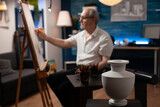 Close up of design of white vase on table with art instruments in artwork studio while elder artist drawing on canvas in background. Aged man working on authentic masterpiece at home
