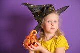 Little girl in witch costume for Halloween with ceramic pumpkin Jack on Lilac background