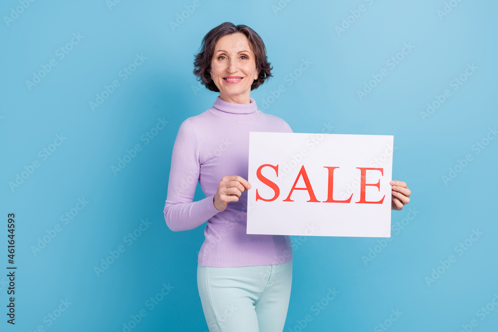 Photo of mature woman happy positive smile hold banner poster advertising sale isolated over blue color background Photo of mature woman happy positive smile hold banner poster advertising sale isolated over blue color background