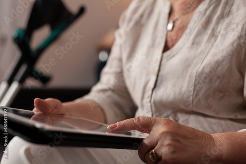 Close up of unrecognisable aged hands touching tablet screen at home. Elder woman using digital device while sitting on living room couch with crutches for walking. Senior adult with technology
