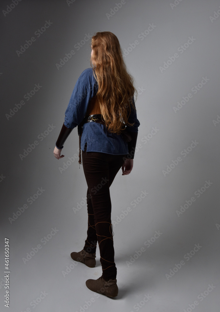 Full length portrait of red haired woman wearing medieval viking inspired costume, standing pose with back to the camera, against studio background. Full length portrait of red haired woman wearing medieval viking inspired costume, standing pose with back to the camera, against studio background.