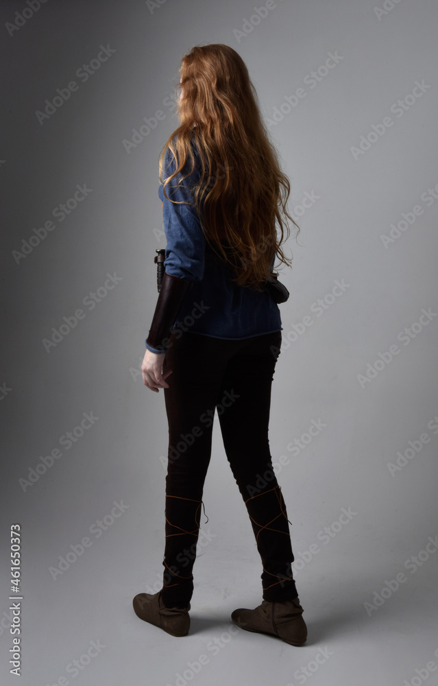 Full length portrait of red haired woman wearing medieval viking inspired costume, standing pose with back to the camera, against studio background. Full length portrait of red haired woman wearing medieval viking inspired costume, standing pose with back to the camera, against studio background.