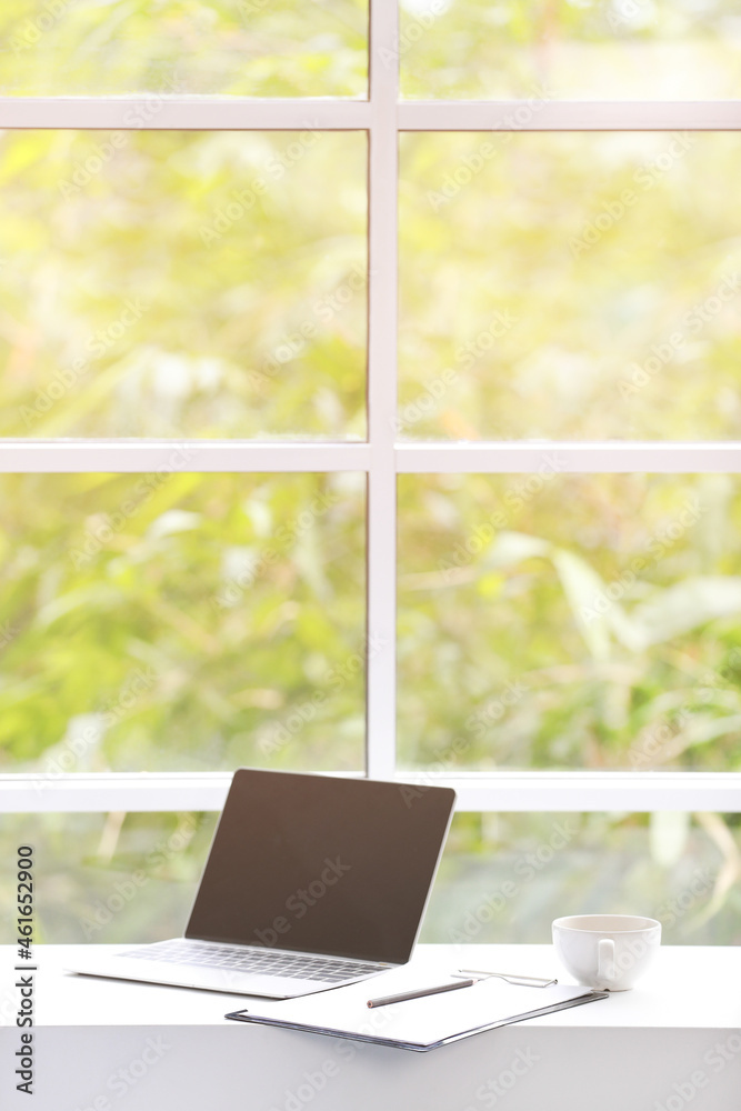 Blank black screen laptop computer for text copy space advertisement place on white table counter with paper notebook coffee cup in home office near glass windows with tree garden view in warm light Blank black screen laptop computer for text copy space advertisement place on white table counter with paper notebook coffee cup in home office near glass windows with tree garden view in warm light