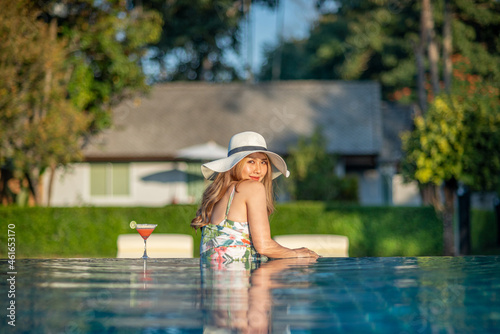 A beautiful young Asian woman with long hair wearing a white straw hat and a glass of cocktail on the edge of a swimming pool on a sunny day. Tropical summer vacation and holiday concept © tai