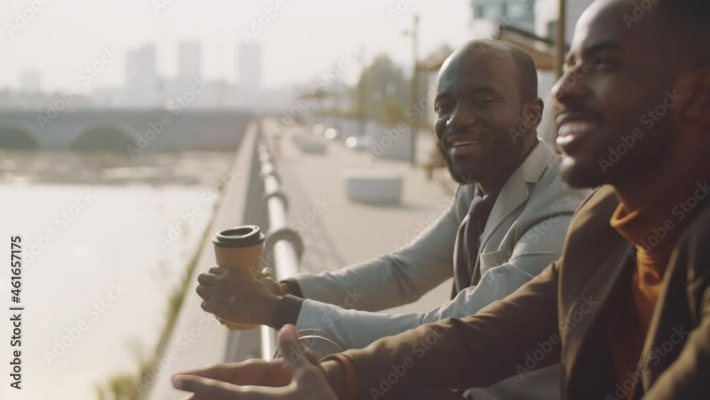 Two cheerful African American businessmen in formalwear leaning on embankment railing and chatting over coffee on summer day
