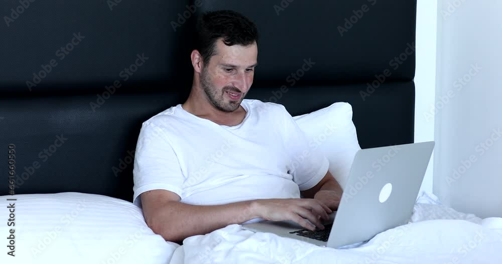 Handsome man working or study on his laptop while sitting on a bed during the Covid-19 pandemic. Young man freelancer working online lying on bed uses laptop. Handsome man working or study on his laptop while sitting on a bed during the Covid-19 pandemic. Young man freelancer working online lying on bed uses laptop.