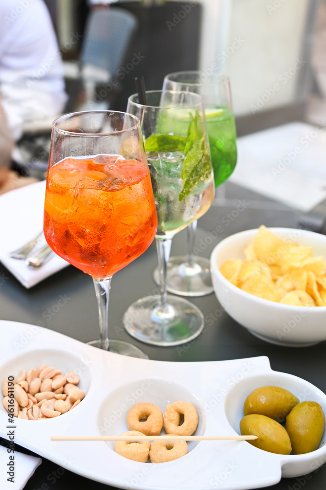 There are three glasses on the table with different colorful cocktails, surrounded by a variety of snacks