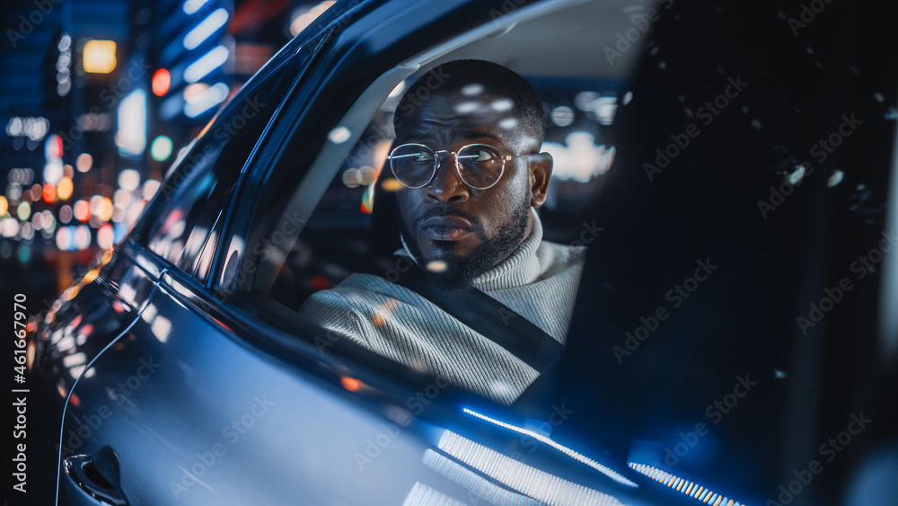 Stylish Black Man in Glasses is Commuting Home in a Backseat of a Taxi at Night. Handsome Male Passenger Looking Out of Window while in a Car in Urban City Street with Working Neon Signs.