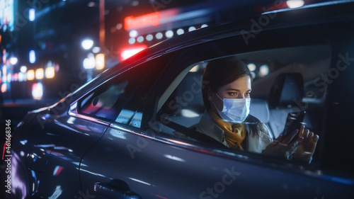 Female Wearing Face Mask is Commuting Home in Backseat of a Taxi at Night. Beautiful Passenger Using Smartphone while in a Car in Urban City Street with Working Neon Signs.