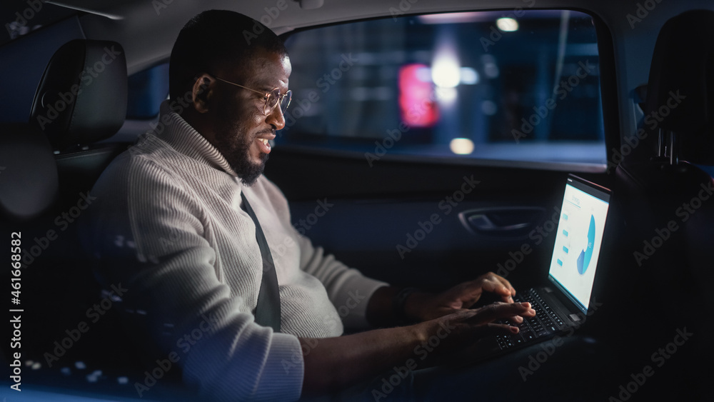 Stylish Black Man in Glasses is Commuting Home in a Backseat of a Taxi at Night. Male Using Laptop Computer and Looking Out of Window while in a Car in Urban City Street with Working Neon Signs. Stylish Black Man in Glasses is Commuting Home in a Backseat of a Taxi at Night. Male Using Laptop Computer and Looking Out of Window while in a Car in Urban City Street with Working Neon Signs.
