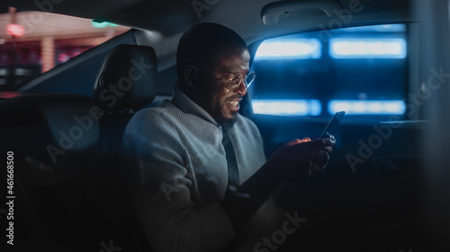 Happy Black Man in Glasses is Commuting Home in a Backseat of Taxi at Night. Handsome Male Using Smartphone and Smiling while in a Car in Urban City Street with Working Neon Signs.