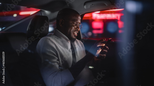 Fotografia Happy Black Man in Glasses is Commuting Home in a Backseat of Taxi at Night