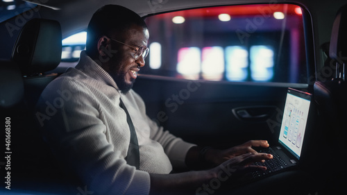 Stylish Black Man in Glasses is Commuting Home in a Backseat of a Taxi at Night. Male Using Laptop Computer and Looking Out of Window while in a Car in Urban City Street with Working Neon Signs. © Gorodenkoff