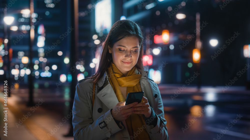 Portrait of a Beautiful Woman in Trench Coat Walking in a Modern City Street with Neon Lights at Night. Attractive Female Using Smartphone and Looking Around the Urban Cinematic Environment.