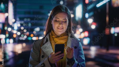 Portrait of a Beautiful Woman in Trench Coat Walking in a Modern City Street with Neon Lights at Night. Attractive Female Using Smartphone and Looking Around the Urban Cinematic Environment. © Gorodenkoff