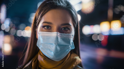 Portrait of a Beautiful Woman Wearing a Protective Face Mask in Trench Coat Standing in a Modern City Street with Neon Lights at Night. Female Looking at Camera in the Urban Cinematic Environment.