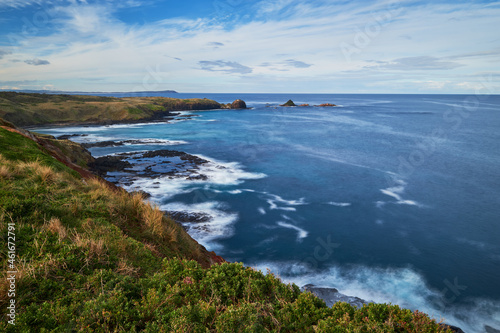 Overlooking a scenic view of a beautiful rocky coastline with waves breaking, deep blue sea in the background and cloudy blue sky on the horizon, photographed at Phillip Island, Australia.
