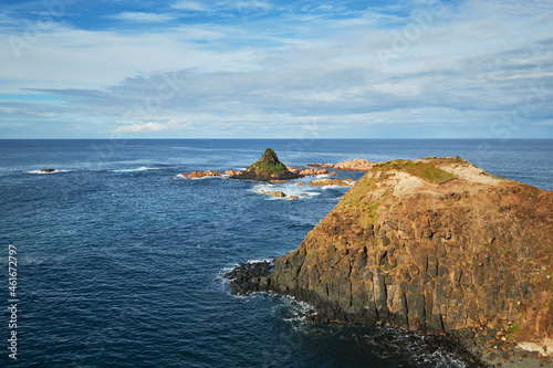 Overlooking a scenic view of a pyramid shaped rocky island with rugged coastline, deep blue sea in the background and cloudy blue sky on the horizon, photographed at Phillip Island, Australia. 