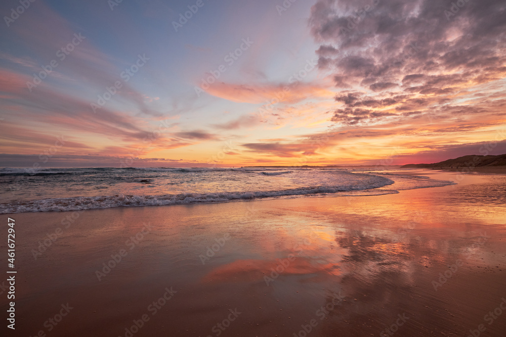 Sunset over a beautiful ocean beach with reflections of clouds in the water, vibrant orange colours illuminate the sky and waves break in the background, in Phillip Island, Australia.
