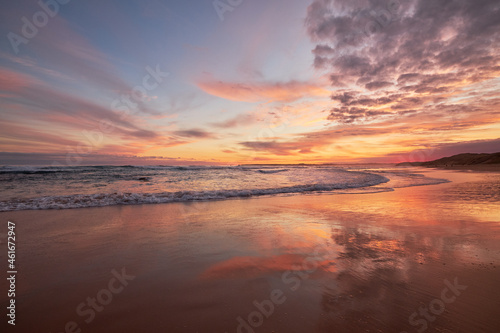 Sunset over a beautiful ocean beach with reflections of clouds in the water, vibrant orange colours illuminate the sky and waves break in the background, in Phillip Island, Australia. 