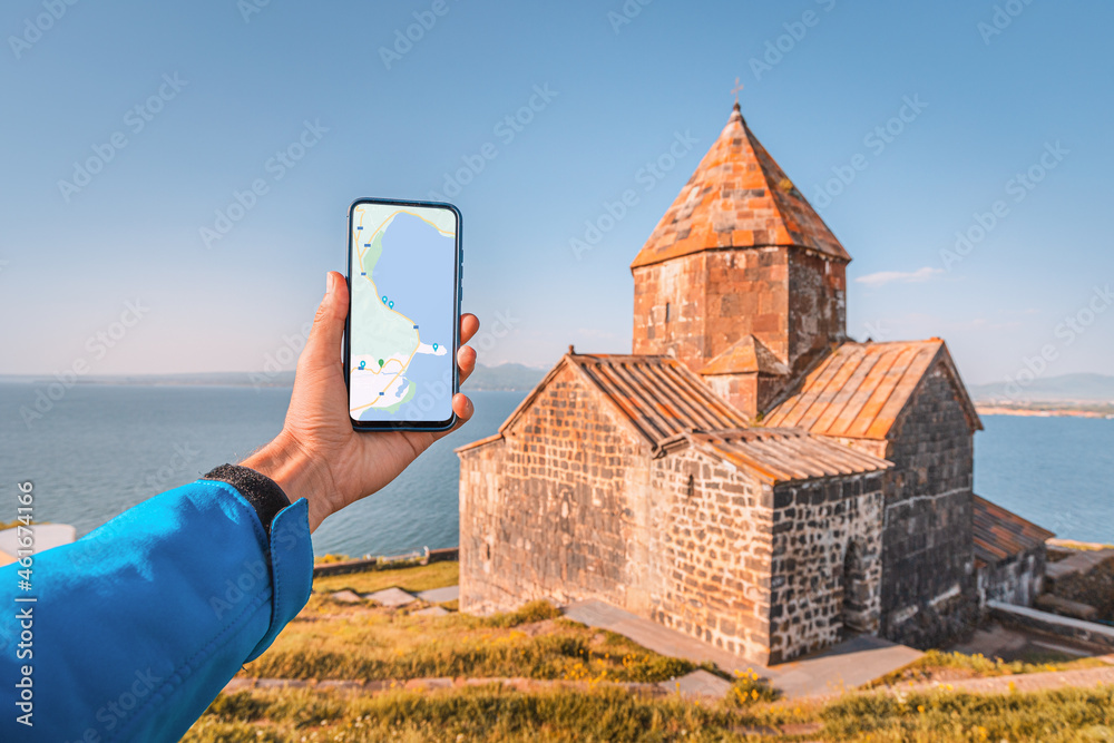 Naklejka premium a hand with phone with maps app for navigating to attractions. At the background - Lake Sevan and the famous Armenian monastery Sevanavank