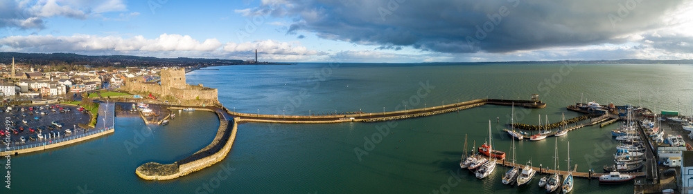 Wide aerial panorama of Carrickfergus near Belfast with medieval Norman castle, harbor, marina, yachts, breakwater, boat ramp, Belfast Laugh, parking lot. Sunset light and strom clouds in winter.