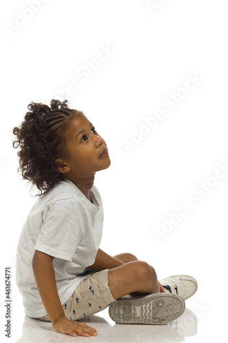 Small black boy in shorts and shirt is sitting on a floor and looking up. Side view. Full length, isolated. photo