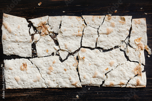 Dry flat bread broken in pieces and divided, with crumbs, on dark wooden background
