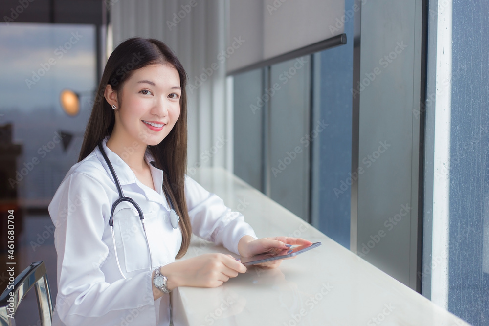 Asian professional woman doctor dressed in white medical coat sits on chair at a hospital library to search some information on a tablet for treatment in health care concept. Asian professional woman doctor dressed in white medical coat sits on chair at a hospital library to search some information on a tablet for treatment in health care concept.