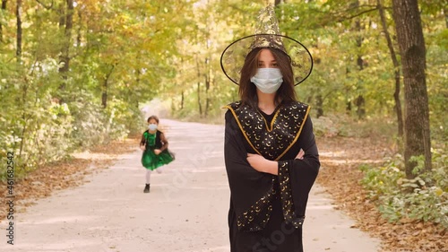 Close up video of a young pretty woman in black dress for the Halloween party in a medical mask walking in the forest and a little girl in a medical mask running by. photo