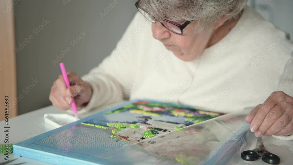Elderly woman with short grey hair in woolen jacket and glasses spends retirement time laying out diamond mosaic carefully closeup Elderly woman with short grey hair in woolen jacket and glasses spends retirement time laying out diamond mosaic carefully closeup