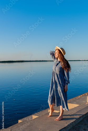 Adult woman in blue dress and straw hat walking near the river. Fortieth people.
