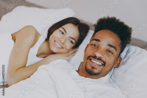 Side view smiling happy young couple two family man woman in casual white clothes lying in bed looking aside rest relax spend time together in bedroom lounge home in own room house wake up good day.