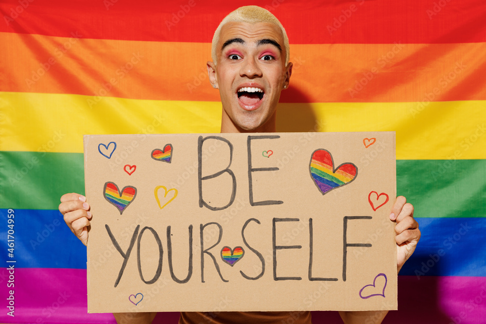 Young queer bisexual surprised blond latin gay man with make up in beige tank shirt hold card sign with be yourself title text on rainbow flag background studio portrait People lgbt lifestyle concept. - obrazy, fototapety, plakaty 