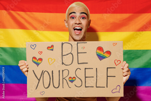 Young queer bisexual surprised blond latin gay man with make up in beige tank shirt hold card sign with be yourself title text on rainbow flag background studio portrait People lgbt lifestyle concept. photo