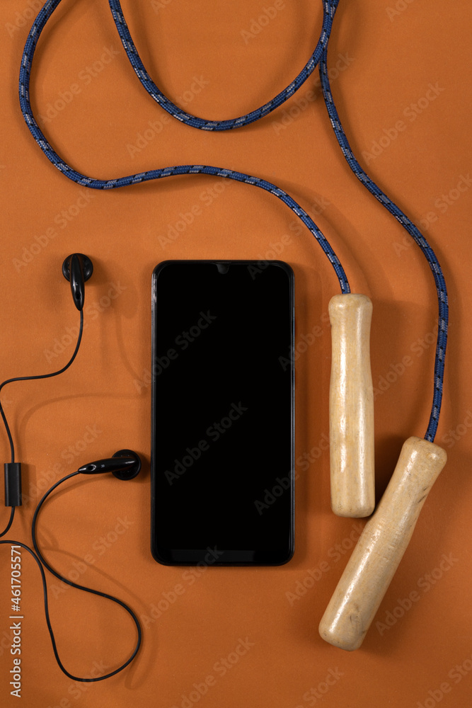 Vertical image of black screen of smartphone,earphones,jump rope on the dark orange background.Empty space Vertical image of black screen of smartphone,earphones,jump rope on the dark orange background.Empty space