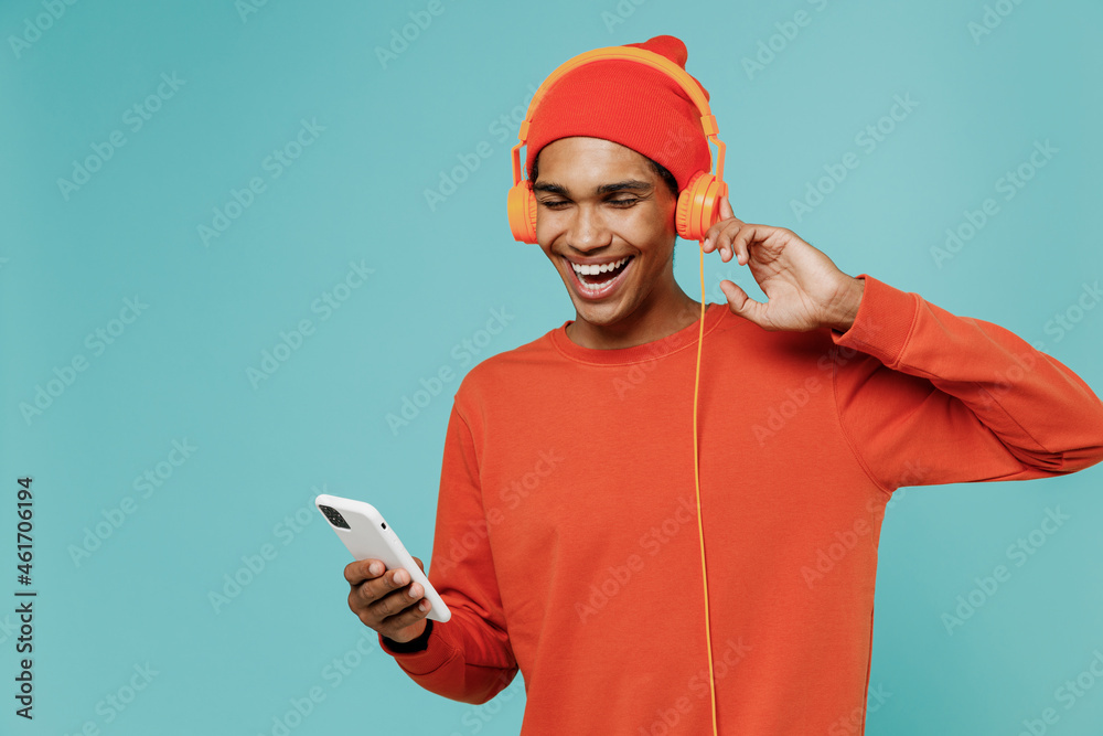 Φωτογραφία Young satisfied smiling fun happy african american man 20s in orange shirt hat headphones use mobile cell phone listen to music dancing isolated on plain pastel light blue background studio portrait