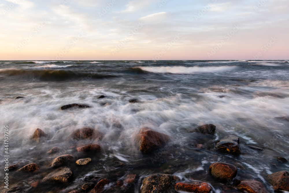 Long exposure photo of waves reaching to rocky coast during dusk. Whitecaps waves are reaching to shore during sunset. Motion of ripples and coastal rocks at sundown