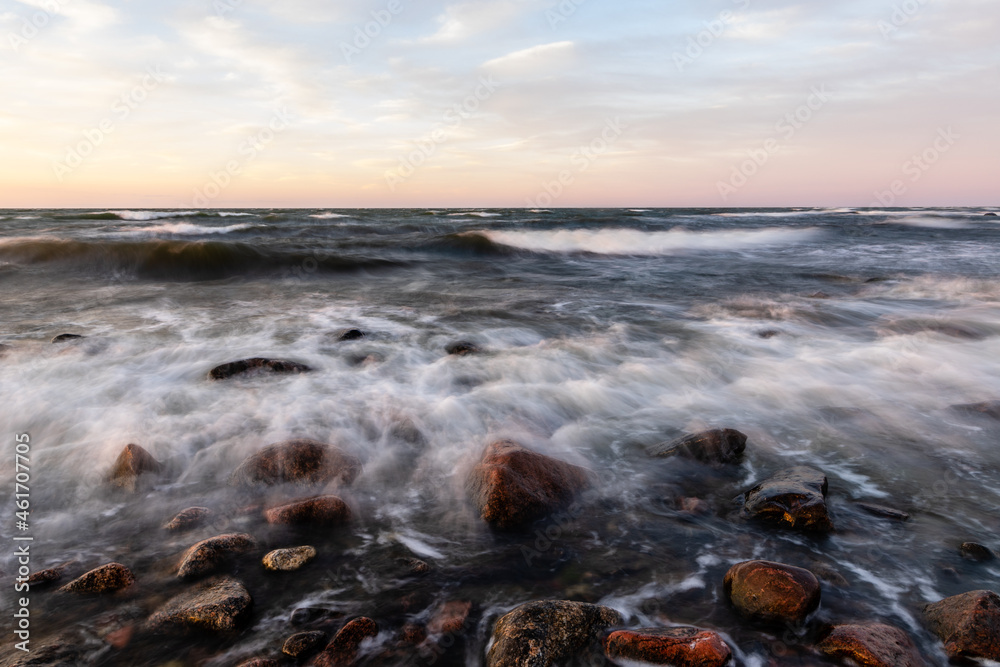 Fototapeta premium Long exposure photo of waves reaching to rocky coast during dusk. Whitecaps waves are reaching to shore during sunset. Motion of ripples and coastal rocks at sundown