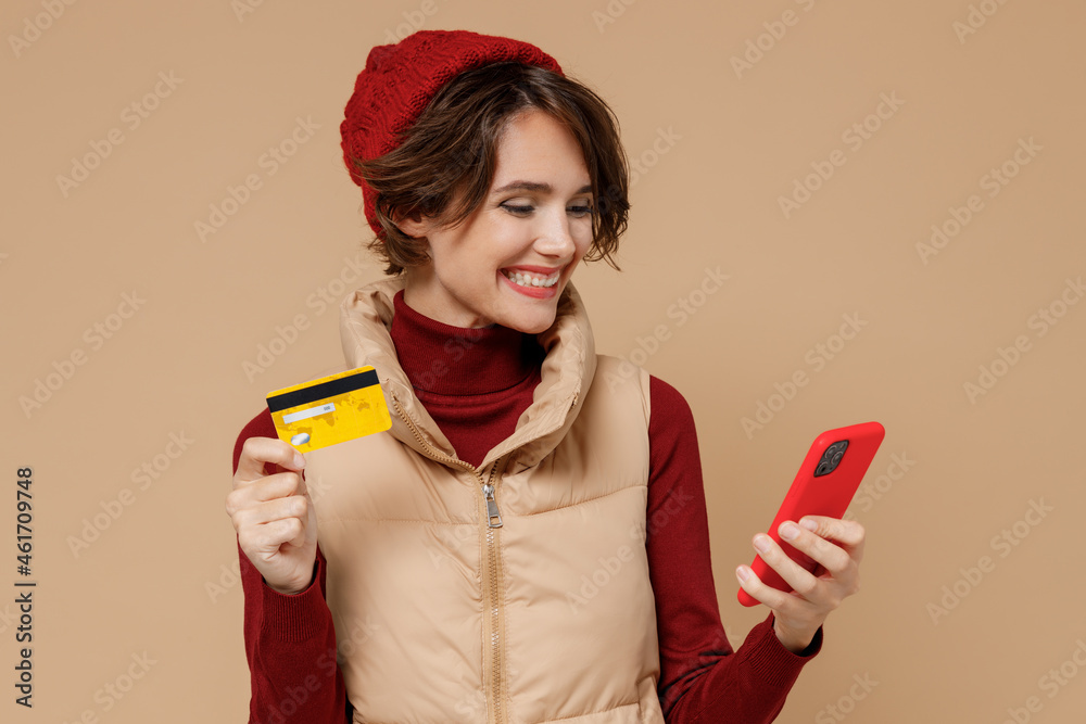 Young woman wears red turtleneck vest beret using mobile cell phone hold credit bank card doing online shopping order delivery booking tour isolated on plain pastel beige background studio portrait Young woman wears red turtleneck vest beret using mobile cell phone hold credit bank card doing online shopping order delivery booking tour isolated on plain pastel beige background studio portrait