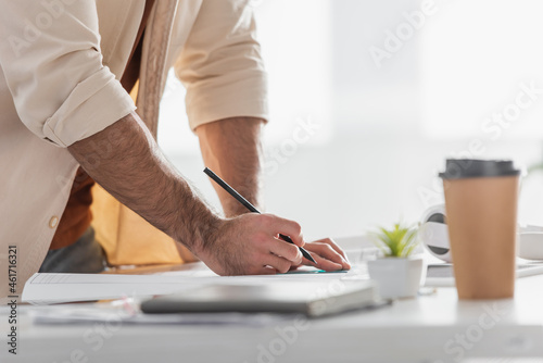 partial view of designer with pencil near blueprint, blurred paper cup and notebook